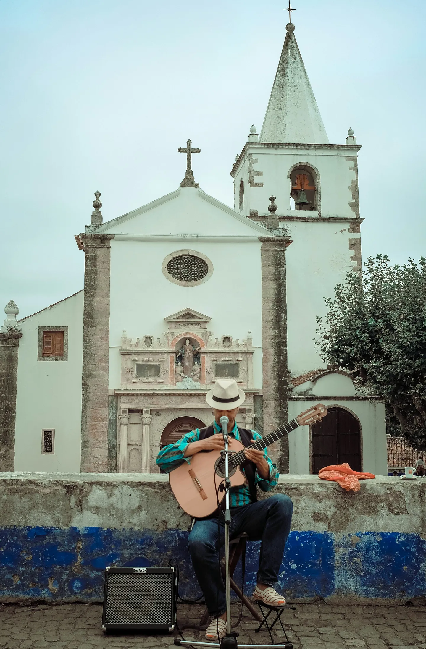 Mann mit Hut, Gitarre, Mikrofon und Lautsprecher sitzt vor einer Kirche und wird gleich Musik machen.